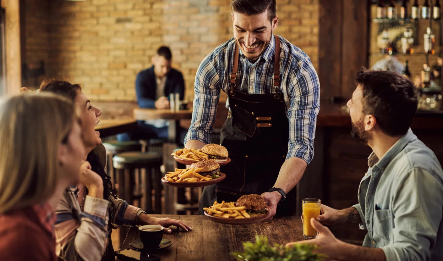Smiling waiter in an apron serving burgers and fries to a group of friends seated at a wooden table inside a cozy restaurant with a brick wall and bar in the background.